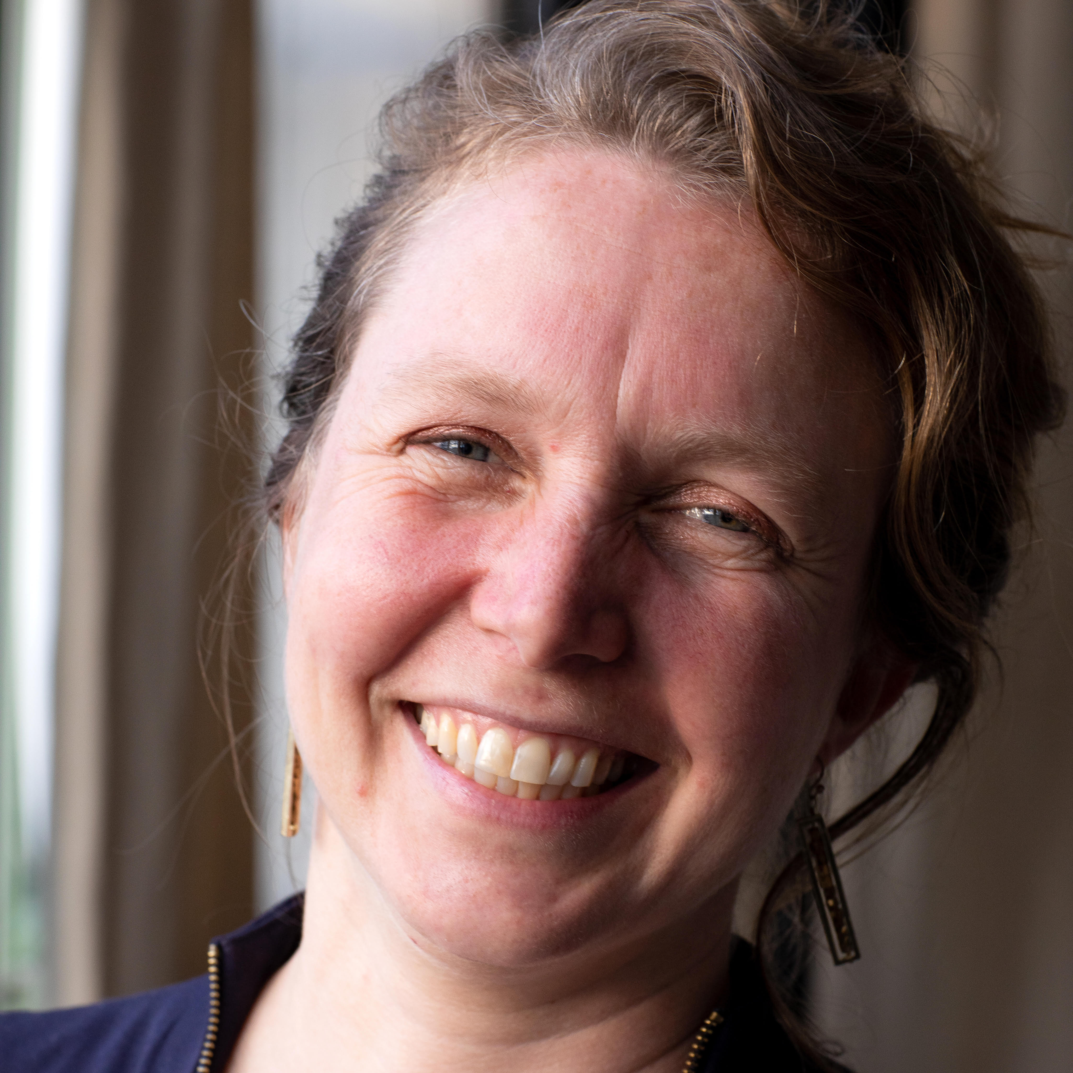 A smiling woman with light brown hair and a dark shirt, standing indoors. She is positioned in front of a soft-focus background with natural light illuminating her face, highlighting her features.