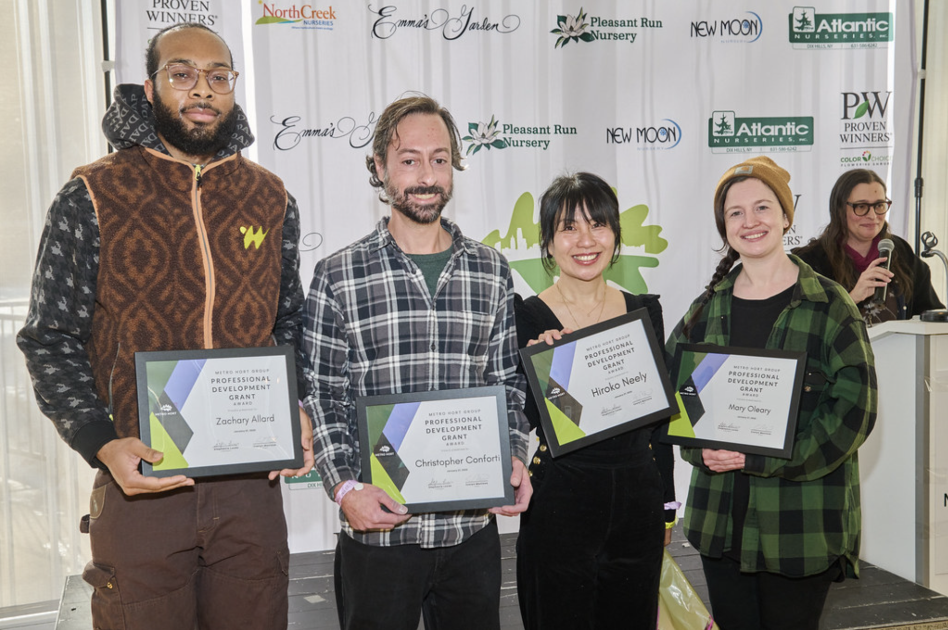 Four individuals stand together holding award plaques at an event. From left to right: Zachary Alford with glasses and a patterned sweater, Christopher Coffin in a plaid shirt, Hilda Neely in a black top, and Mary Cherry in a green plaid shirt. A backdrop features sponsor logos.