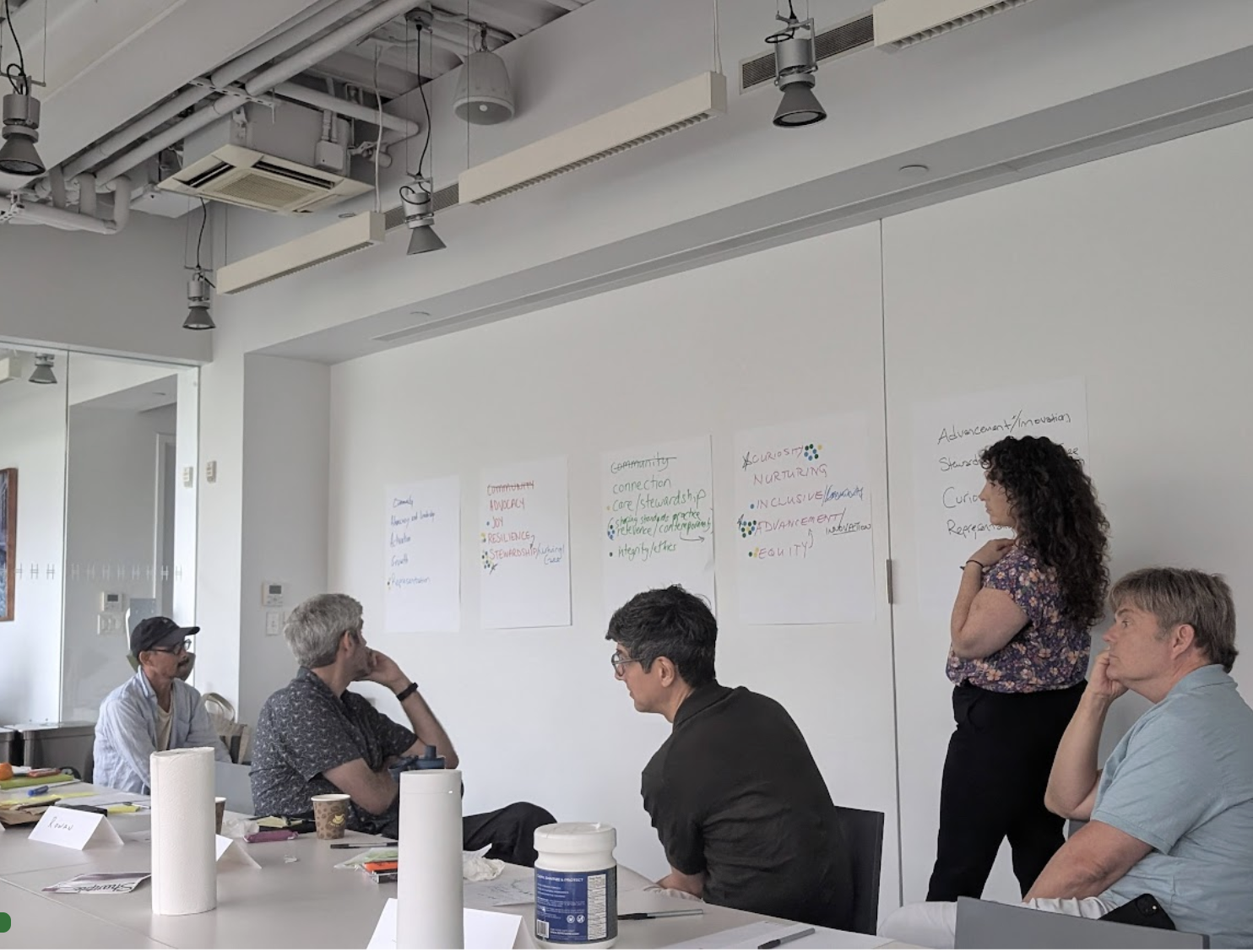 A group of people sitting at a table in a bright, modern conference room. One person stands and presents, while others listen. Various colored notes are taped to the wall, displaying strategic planning points and ideas.