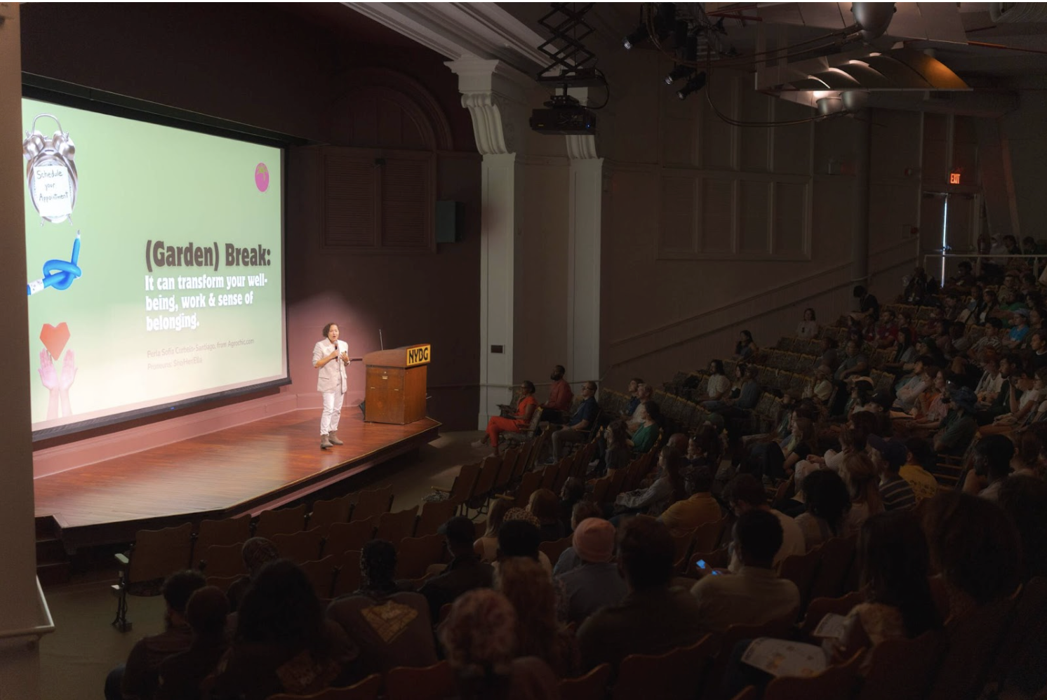 A speaker presenting on a stage in front of an audience in a theater. The screen displays the title '(Garden) Break: It can transform your well-being, your sense of belonging.' Attendees are seated, facing the speaker and the screen.
