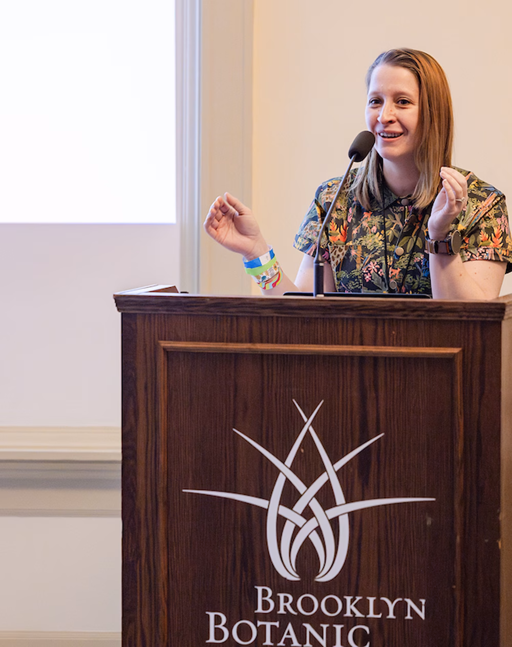 A speaker addressing an audience from a wooden podium marked with the "Brooklyn Botanic" logo. She is wearing a floral-patterned shirt and gesturing with her hands while engaging the audience.