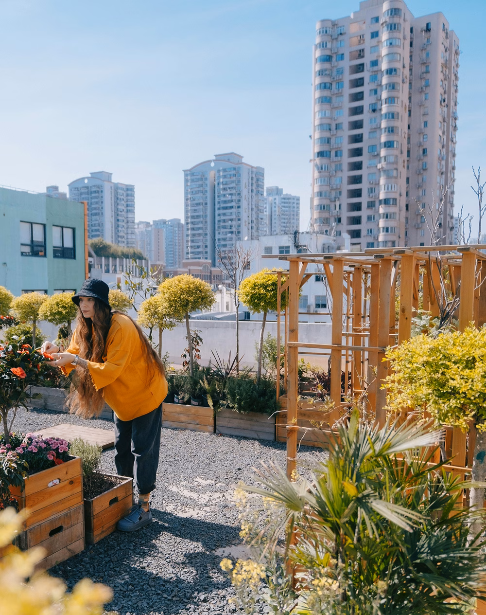 A woman wearing an orange sweater and black hat tending to flowers in a rooftop garden. Surrounding her are neatly arranged wooden planters and greenery, with modern buildings visible in the background under a clear blue sky.