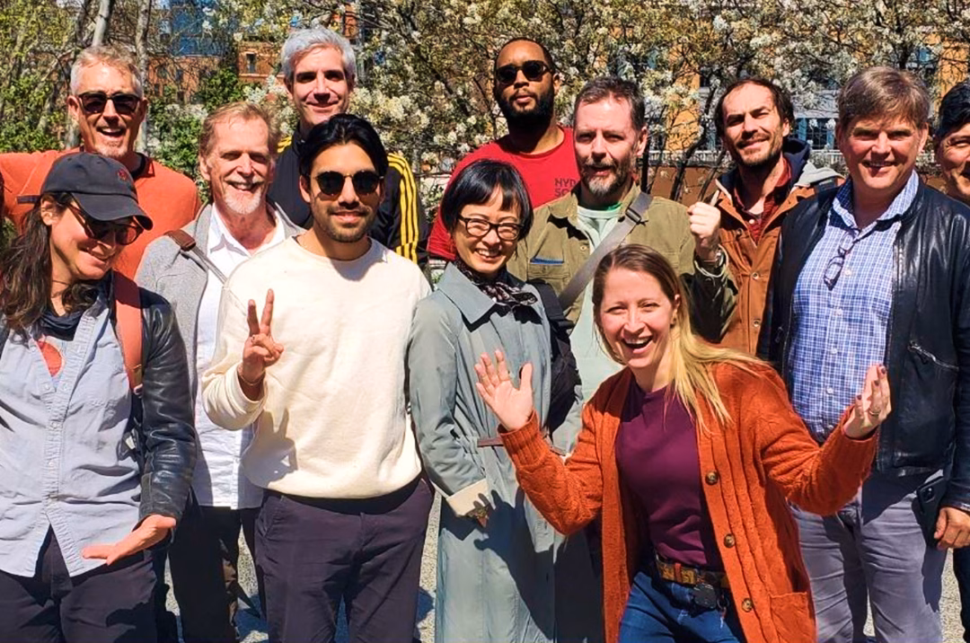A diverse group of thirteen people stands outdoors, smiling and posing for a photo. Flowers bloom in the background, and the group includes men and women of various ages, wearing casual clothing. Some individuals show peace signs and gestures.