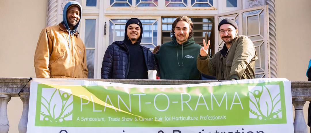 Four men pose in front of a building, standing behind a large banner that reads 'PLANT-O-RAMA: A Symposium, Trade Show & Career Fair for Horticulture Professionals.' The men are smiling and wearing casual clothing suitable for the outdoors.