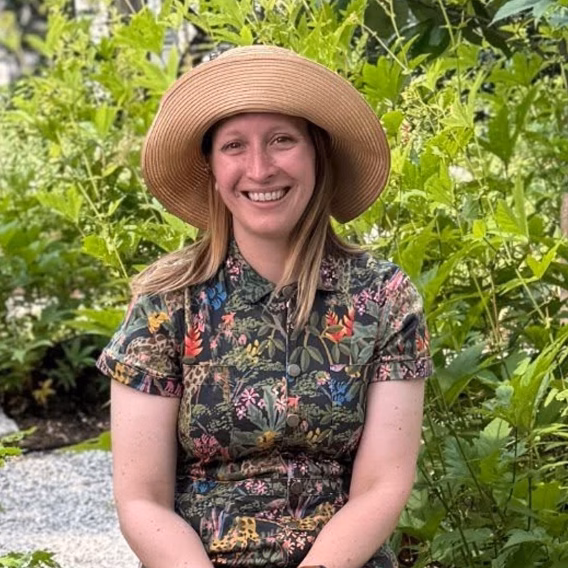 A woman seated outdoors among greenery, wearing a patterned shirt and a wide-brimmed straw hat. She is smiling, with her long hair falling over her shoulders, surrounded by lush plants in a garden setting.