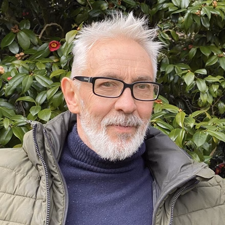 A man with gray hair and a beard wearing glasses and a green jacket stands in front of a background of leafy bushes, showcasing green leaves and some red flowers.