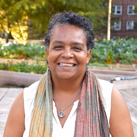 A woman with curly hair, wearing a white top and a multicolored scarf, smiles outdoors in a gardening area. Green plants and raised beds are visible in the background, suggesting a community or urban garden setting.