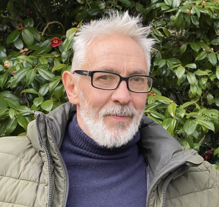 An older man with gray hair and a beard smiles while wearing glasses and a green jacket. He is standing in front of a green shrub with red flowers, which adds a natural backdrop to the image.