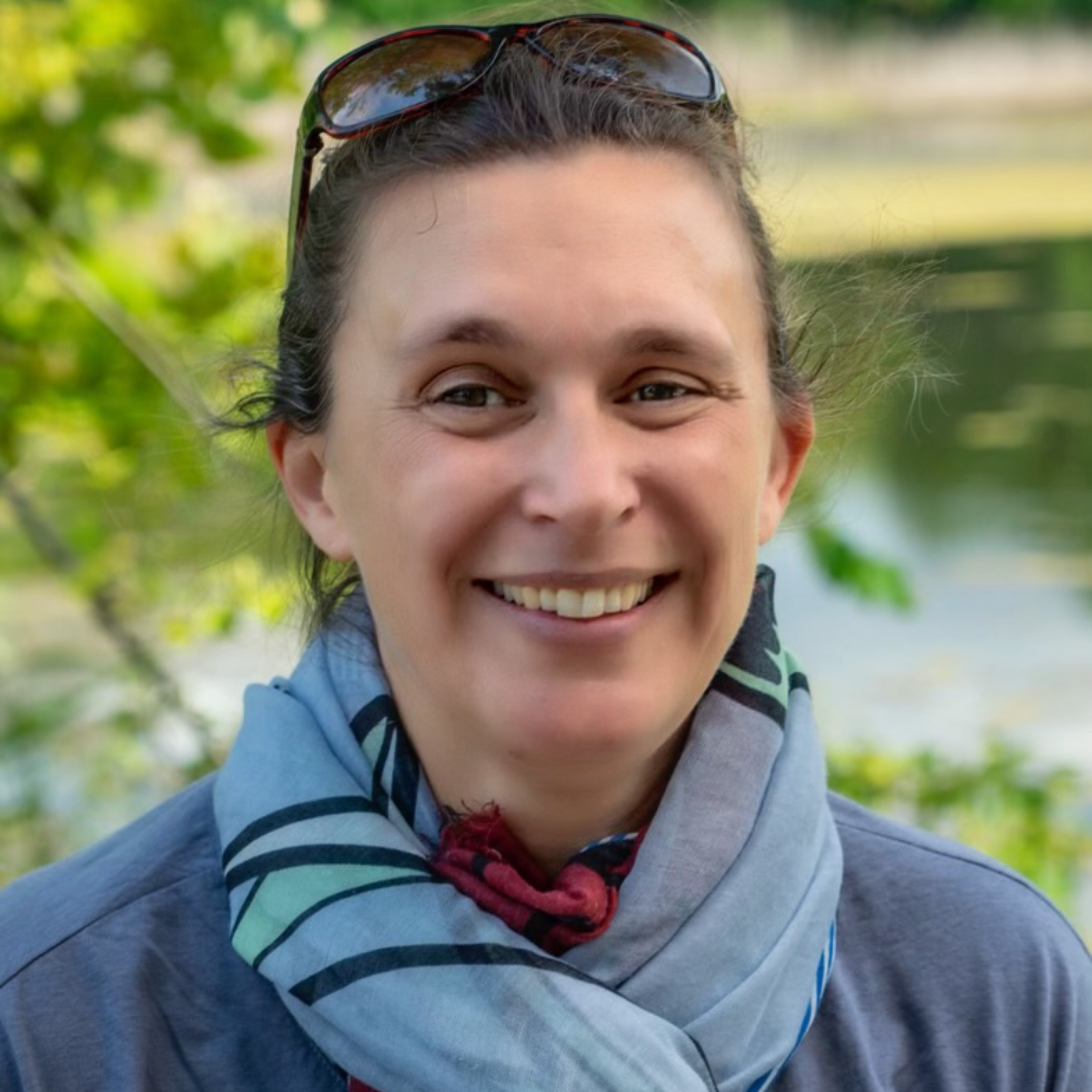 A woman with a pleasant smile, wearing sunglasses on her head and a striped scarf, is standing outdoors near a body of water. Lush greenery surrounds her, indicating a natural setting.