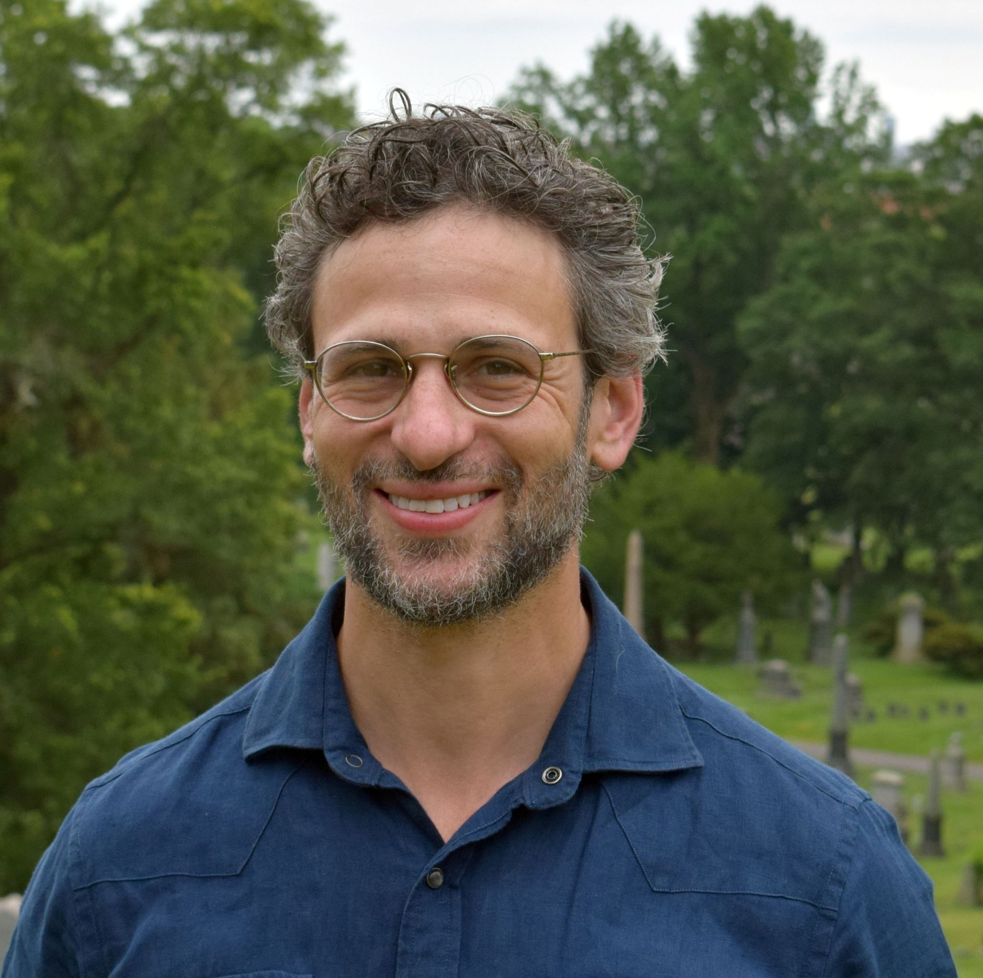 A man with curly hair and glasses smiles while standing outdoors. He wears a blue button-up shirt and is surrounded by trees and gravestones in a cemetery setting.