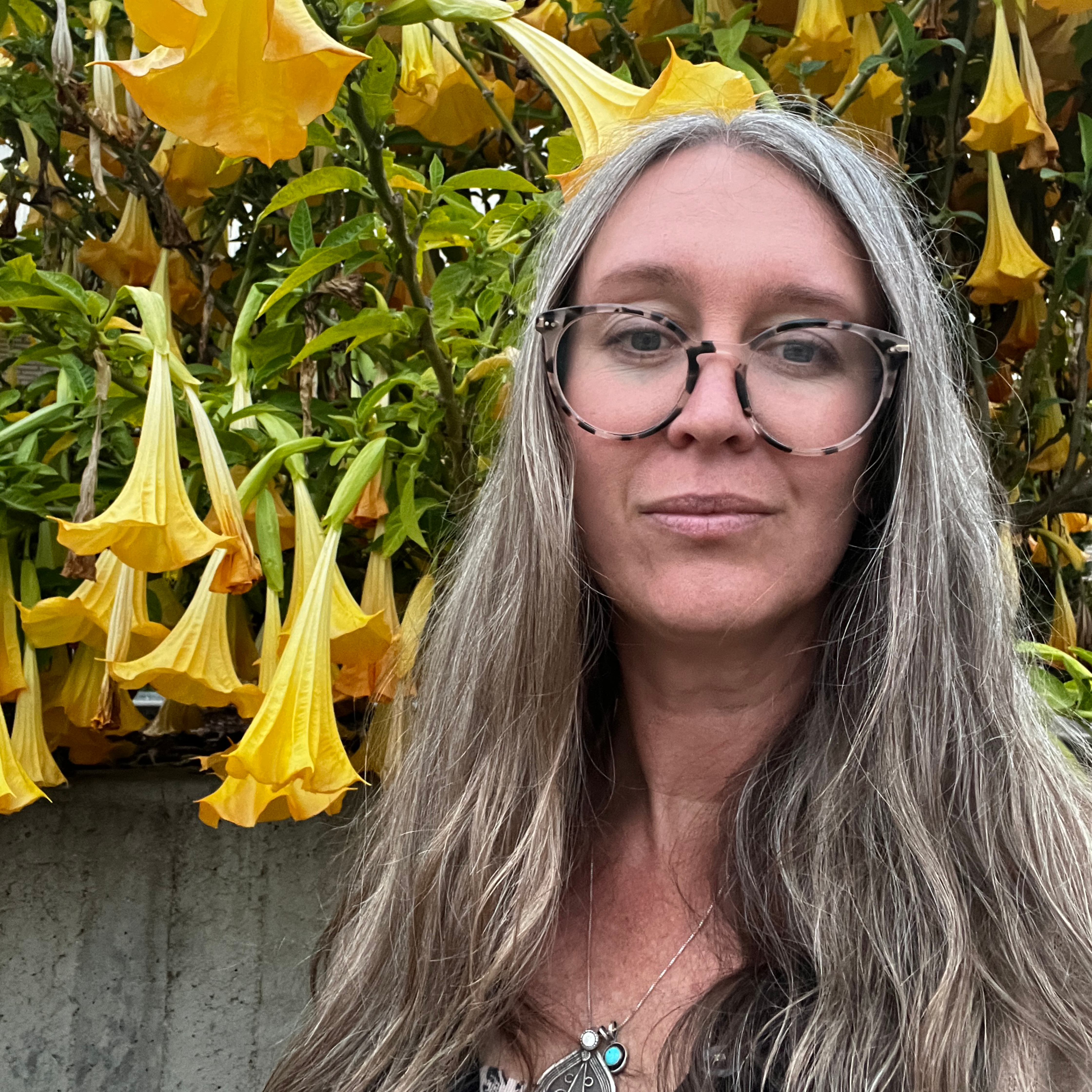 A woman with long, straight gray hair, wearing glasses, stands in front of a vibrant background filled with yellow trumpet-shaped flowers. The flowers are lush and prominent, creating a colorful natural setting.