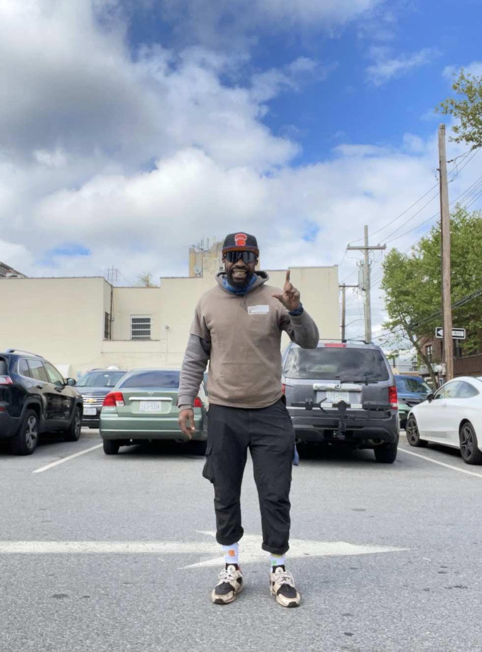 A man stands in a parking lot with a raised index finger, wearing sunglasses, a cap, a long-sleeve gray shirt, and black pants. The background features cars, a building, and a cloudy sky with hints of blue and green foliage.