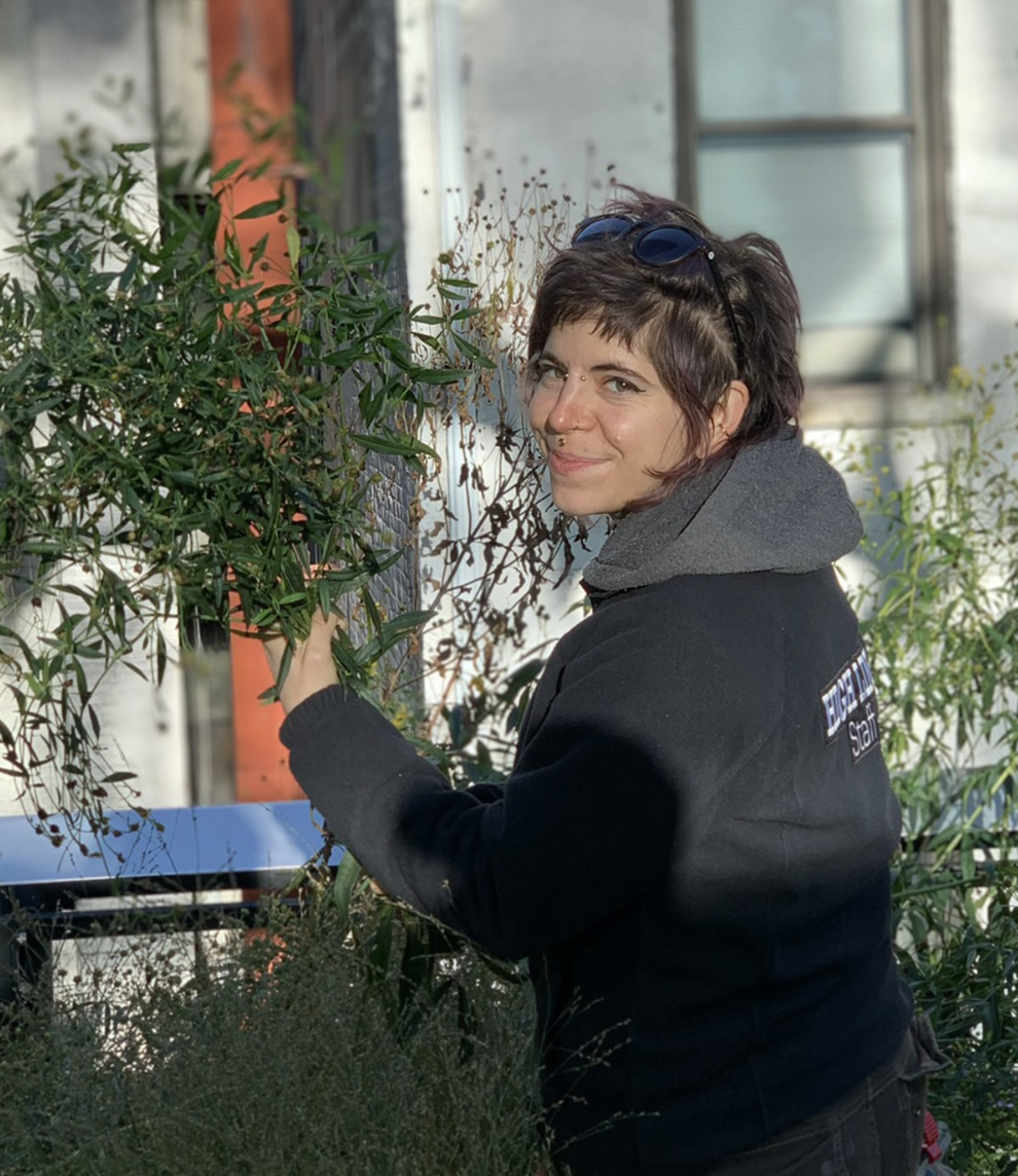A woman with short brown hair, wearing a black jacket, smiles while holding a green plant in a garden. Sunlight highlights her face, and there are various plants in the background, contributing to a natural setting.
