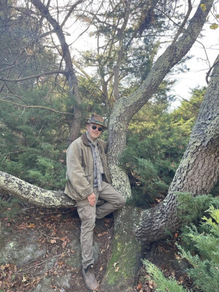 A man wearing a hat, sunglasses, and a light jacket is sitting on a large branch of a tree, surrounded by green foliage. He is posed casually, with one leg resting on the branch, in a natural outdoor setting.
