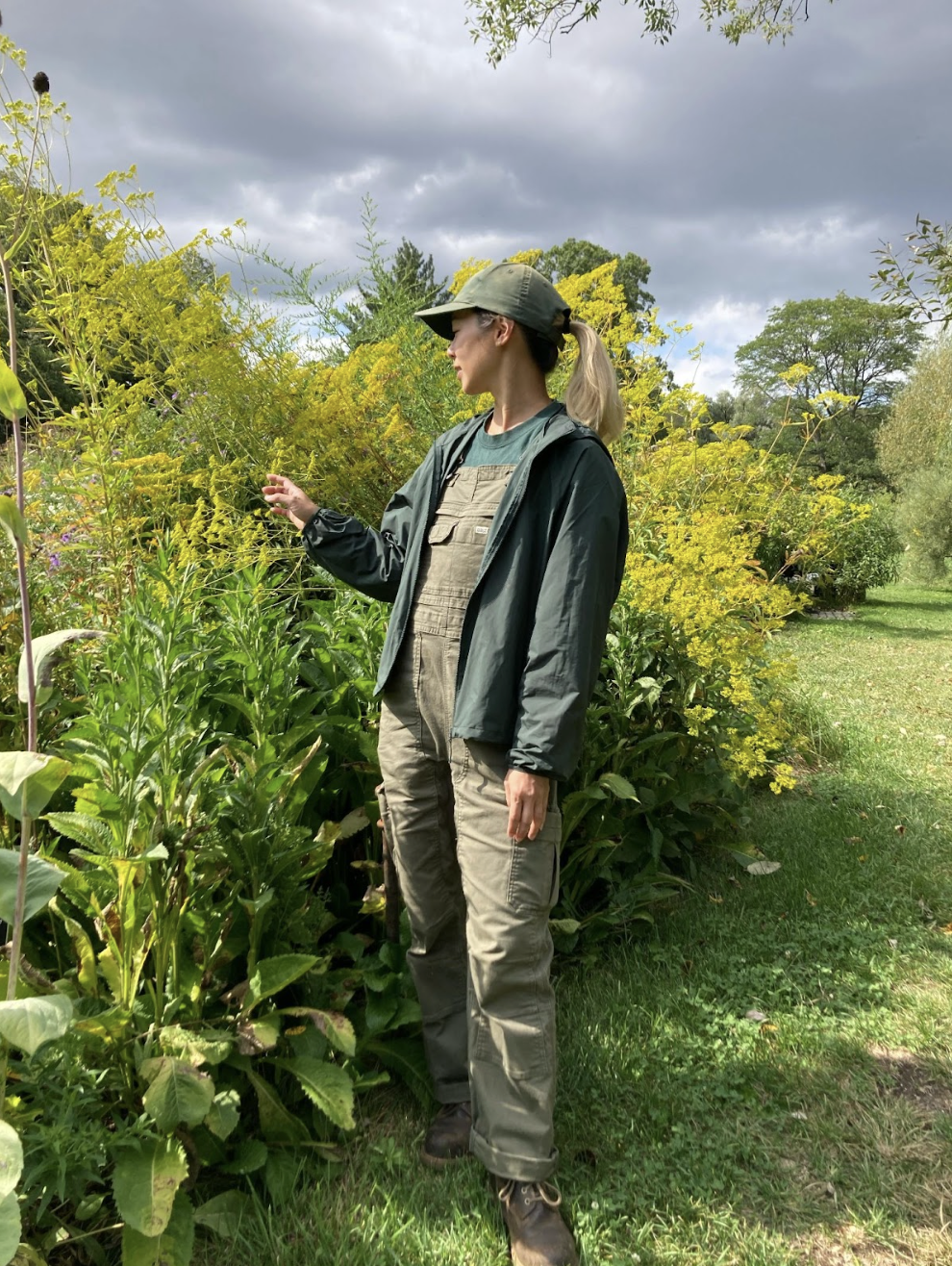 A woman wearing a green jacket and olive overalls stands in a garden filled with yellow flowers. She is looking at a flower while surrounded by lush greenery, under a cloudy sky.
