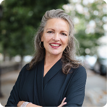A woman with long, gray hair wearing a black blouse stands outdoors, smiling confidently. She is in front of blurred greenery and a city street, with her arms crossed. The image presents a professional and approachable demeanor.