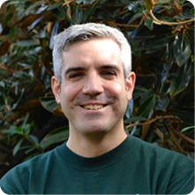 A man with short gray hair and a slight smile stands in front of greenery. He is wearing a dark green sweatshirt and appears to be outdoors, surrounded by leaves.