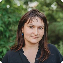 A woman with shoulder-length brown hair and earrings stands outdoors against a background of green foliage. She wears a black button-up shirt and appears to be looking directly at the camera.