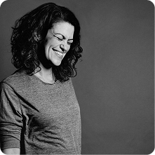 A woman with curly hair, wearing a gray top, smiling slightly while looking down. The image is in black and white and features a simple gray background, emphasizing her expression and form.