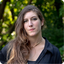 A woman with long, wavy brown hair standing outdoors. She is wearing a black shirt and has a neutral expression. The background features green foliage, indicating a natural setting.