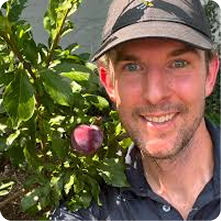 A man smiling while posing next to a fruit tree. He is wearing a dark cap and a shirt, with a visible red apple hanging from the tree branch on the left. The background features green leaves, indicating a garden or park setting.