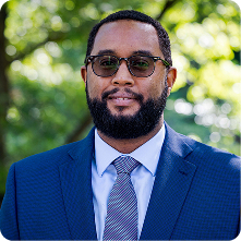 A man wearing glasses and a suit, standing outdoors with blurred greenery in the background. He has a short beard and is looking directly at the camera with a neutral expression.