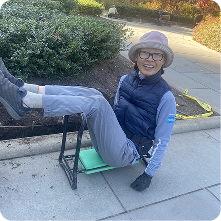 An individual sitting on a portable stool with legs bent and feet elevated. They are wearing a blue long-sleeve shirt, a dark vest, and gloves, and have a light-colored hat. The background shows landscaped greenery and a paved walkway.