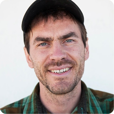 A man with short dark hair and a beard smiles directly at the camera. He wears a plaid shirt and a black cap, with a neutral background. The image conveys a friendly demeanor, suitable for an About Us page.