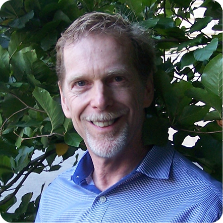 A man with short, light brown hair and a beard smiles at the camera, standing outdoors in front of leafy green foliage. He is wearing a blue checkered shirt. The background includes various shades of green leaves.