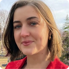 A woman with shoulder-length brown hair smiles softly, wearing a red jacket. She is outdoors with trees and a cloudy sky in the background, suggesting a calm and natural setting.
