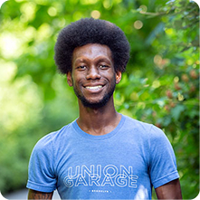 A smiling man with an afro hairstyle stands outdoors among green foliage, wearing a blue t-shirt with the text "UNION GARAGE" printed on it. The background features blurred leaves, indicating a natural setting.