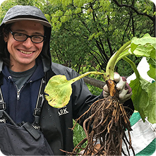 A person wearing glasses, a hoodie, and a rain jacket smiles while holding up a large root vegetable with green leaves. The background includes trees and greenery, indicating an outdoor setting.