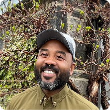 A man with a beard and mustache smiles while standing outdoors. He wears a gray baseball cap and a green button-up shirt. Behind him, there are green leaves and the texture of a stone wall, creating a natural backdrop.