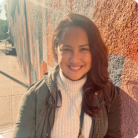 A woman sitting outdoors in front of a colorful mural. She has long brown hair and is wearing a light sweater and a jacket. The sunlight creates a warm glow on her face as she smiles at the camera.