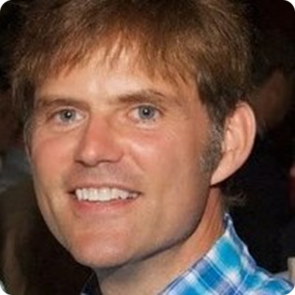 A smiling man with short brown hair and blue eyes, wearing a blue and white checkered shirt. The background is blurred, focusing on his face and expression.