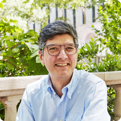 A man with salt-and-pepper hair wearing glasses smiles while sitting outdoors in front of lush green foliage. He is dressed in a light blue button-up shirt, with sunlight filtering through the leaves behind him.