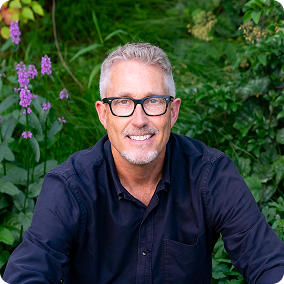 A middle-aged man with glasses is seated outdoors among green foliage and purple flowers. He is smiling at the camera, wearing a black button-up shirt, with a blurred natural background enhancing the image.