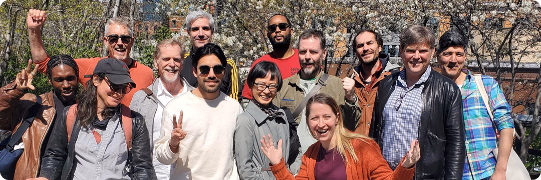 A diverse group of 15 people smiling and posing for a photo outdoors. They are standing in front of trees with green leaves and some flowers. Several individuals are making peace signs and gestures of celebration.