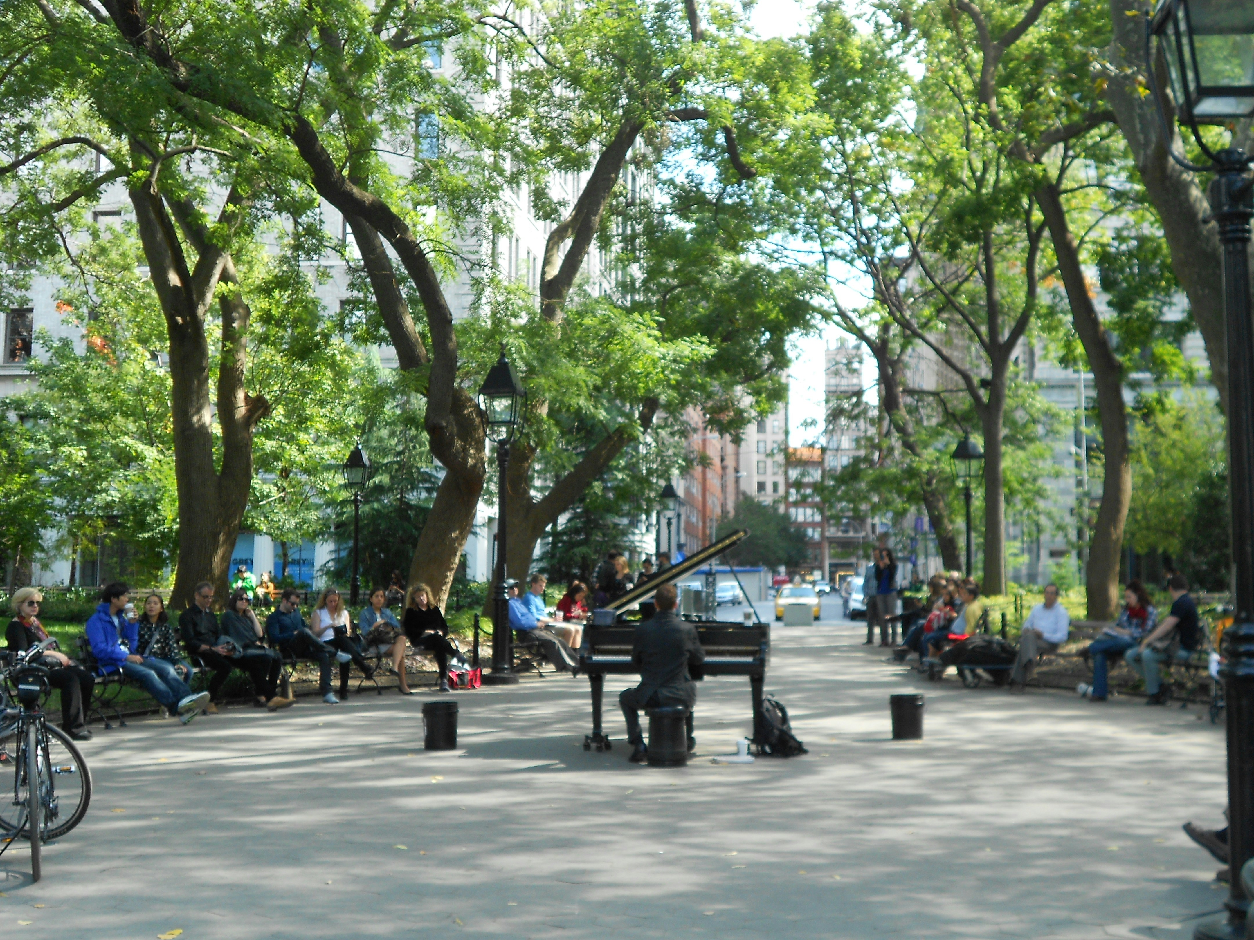 A street musician playing a piano in a green park lined with trees. Several people are seated on benches, listening to the performance. Bicycles are parked nearby, and buildings are visible in the background.